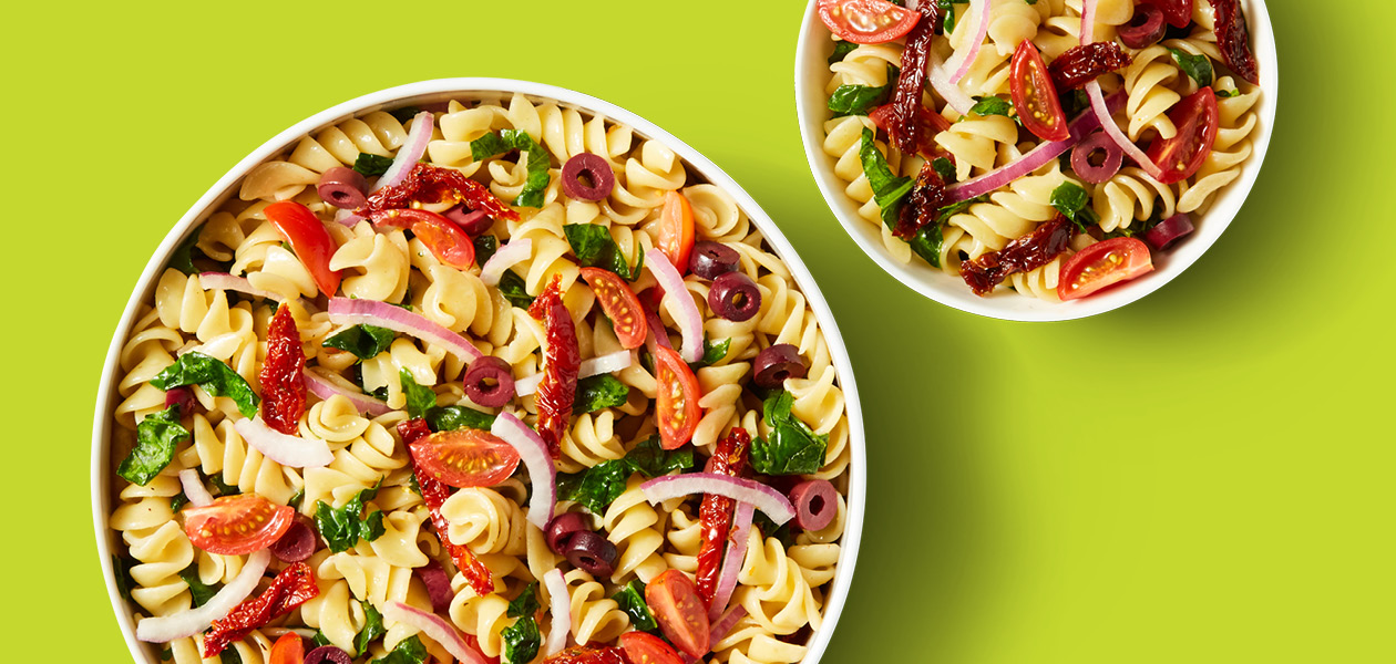 Two bowls of colourful pasta salad featuring rotini pasta, red onions, cherry tonatoes, sundried tomatoes, spinach, and kalamata olives.